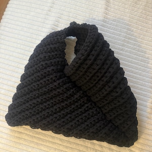 May include: A black, handmade crochet bag with a unique, open design. The bag is made of thick, textured yarn, and the stitches create a ribbed pattern. The bag's shape is triangular, with a handle that forms a loop.