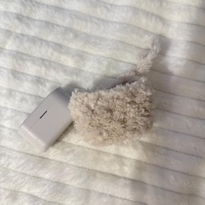 May include: A light beige, fluffy, rectangular pouch with a matching cord, next to a white rectangular power adapter. The items are on a white, textured, plush surface. The pouch appears to be made of a soft, fuzzy material.
