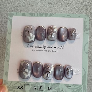 Pode incluir: Um conjunto de dez unhas postiças com acabamento olho de gato em tom malva, apresentando detalhes florais brancos e toques metálicos prateados. O cartão inclui o texto One beauty one world e one anmoun and one heart, com tamanhos XS, S, M e L.