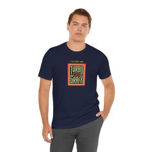 May include: A navy blue short-sleeve t-shirt featuring a graphic print on the chest. The design includes the yellow text "I'm this old" above a rectangular logo with the words "TURBO GRAFX" and the number "16" in a red and yellow frame.