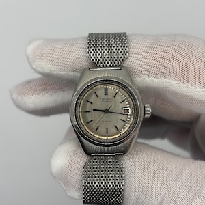 Puede incluir: Un reloj Seiko Automatic vintage con caja plateada y correa de malla. La esfera del reloj es plateada con marcadores de hora y agujas negras. El reloj tiene una ventana de fecha a las 3 en punto.