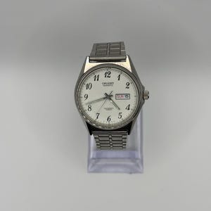 Può includere: Un orologio da polso Orient Quartz argentato con quadrante bianco. L'orologio ha un cinturino in metallo e mostra il giorno e la data. Il quadrante presenta numeri e lancette neri, con il giorno e la data visualizzati alle 3.