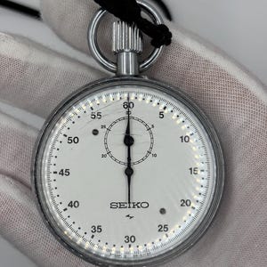 Könnte beinhalten: Eine silberne Seiko Stoppuhr mit weißem Zifferblatt und schwarzen Markierungen. Die Stoppuhr hat eine schwarze Kordel und einen Metallring oben. Das Zifferblatt zeigt Zahlen von 0 bis 60, mit dem Seiko-Logo in der Mitte.