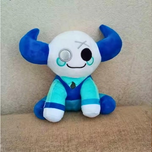 Könnte beinhalten: Ein Plüschtier in sitzender Position, mit einem weißen Kopf und blauen Hörnern. Es hat ein schwarzes Auge und ein Monokel, mit einem türkisblauen und blauen Outfit. Das Spielzeug befindet sich auf einer neutralfarbenen Oberfläche.