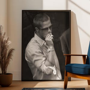 Könnte beinhalten: Gerahmtes Schwarz-Weiß-Foto eines rauchenden Mannes. Der Raum umfasst einen blauen Teppich, einen blauen Stuhl, eine Topfpflanze und ein Fenster. Die Szene wird von natürlichem Licht erhellt.