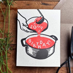 May include: A greeting card with the text "A Sour Birthday" in red, depicting a hand pouring red liquid from a bowl into a pot. A brown envelope and black scissors are next to the card on a wooden surface. Green stems with white flowers are on the left.