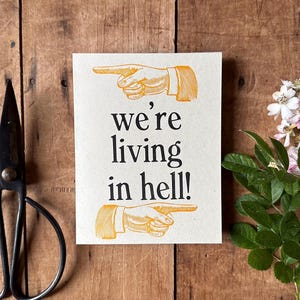 May include: A white card with the text "we're living in hell!" printed in black. Two yellow hands with pointing fingers are on either side of the text. The card is on a wooden surface.