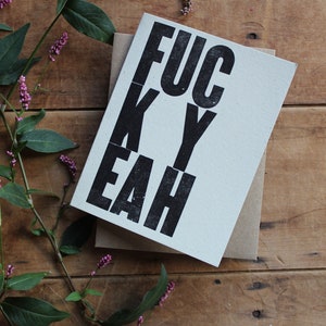 May include: A white card with black lettering that reads "FUCK YEAH" on a wooden surface with green leaves and pink flowers.