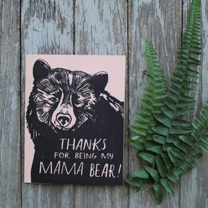 May include: A pink card with a black and white illustration of a bear's face. The text on the card reads "Thanks for being my Mama Bear!"