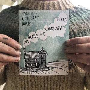 May include: A rectangular greeting card with a black and white illustration of a house with a smoking chimney. The card has the text "ON THE COLDEST DAYS WE BUILD THE WARMEST FIRES" in black lettering against a blue and white background.