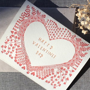 May include: A white card with a red heart design and the text "Happy Valentines Day". The card is surrounded by a border of small red hearts and floral designs.