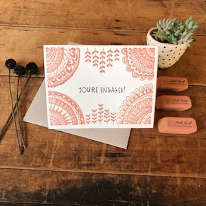 May include: A white card with a pink and white mandala design and the text "You're Engaged!" printed on it. The card is sitting on a brown wooden surface with a small potted succulent plant and three pink erasers with the text "Pink Pearl" printed on them.
