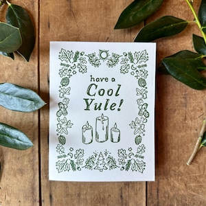 May include: A white card with a green border featuring a festive holiday greeting that reads "have a Cool Yule!" with three candles in the center. The border is decorated with a variety of green illustrations including pine trees, stars, and acorns.