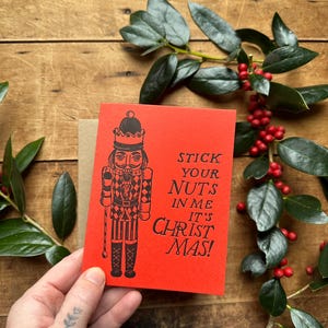 May include: A red Christmas card with a black illustration of a nutcracker and the text "STICK YOUR NUTS IN ME IT'S CHRISTMAS!". A brown envelope is behind the card. Green leaves and red berries are in the background.