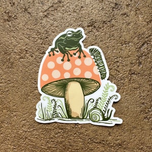 May include: A green frog sits on top of a red and white mushroom with white polka dots. A green caterpillar crawls up the side of the mushroom. A snail sits in the grass below the mushroom.
