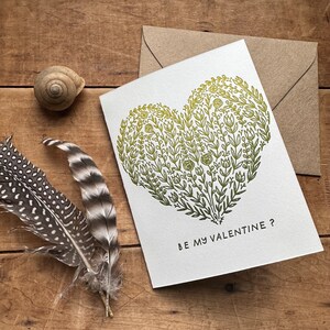 May include: A white card with a green and yellow floral heart design. The text "BE MY VALENTINE?" is printed below the heart.