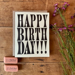 May include: A white card with a yellow patterned background and black text that reads "HAPPY BIRTHDAY!!!". The card is sitting on a wooden surface with two pink erasers.