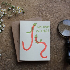 May include: A greeting card with the words "Worm Wishes" and a whimsical illustration of two red worms with green accents. The card is accompanied by a brown envelope, a sprig of white flowers, and a magnifying glass.
