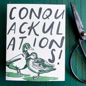 May include: A white card with the text "CONQUACKULATIONS!" in black lettering. Two green ducks are illustrated on the card.