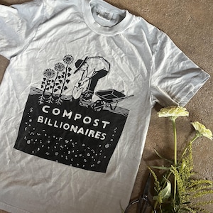 May include: A light gray short-sleeve t-shirt featuring a black graphic print of a person gardening with a shovel and wheelbarrow near sunflowers. The text COMPOST BILLIONAIRES is printed in white within a black rectangular block below the garden scene.