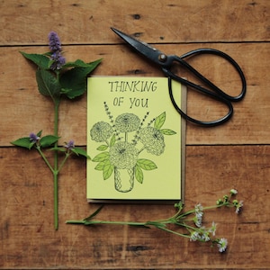 May include: A greeting card with the text "Thinking of You" and a floral illustration, along with a pair of black scissors and sprigs of greenery, all arranged on a wooden surface.