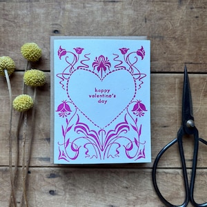 May include: A Valentine's Day card with a light blue background and a pink floral design. The card features a heart shape with the words "happy valentine's day" inside. A pair of black scissors and dried yellow flowers are next to the card.