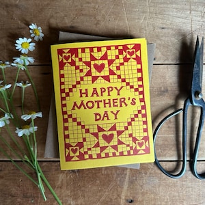May include: A yellow "Happy Mother's Day" greeting card with a red geometric design, a bouquet of white daisies, and a pair of vintage scissors on a wooden surface.
