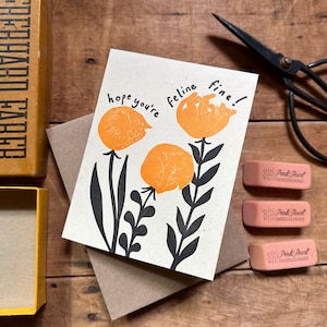 May include: A greeting card with the text "hope you're feline fine!" printed in black. The card features three orange flowers with black stems and leaves. Each flower has a cat sleeping inside.