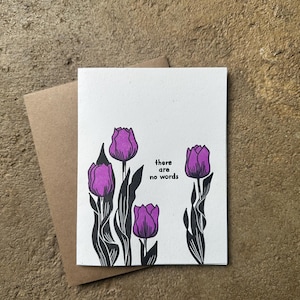 May include: A white greeting card with purple tulip illustrations and the text "there are no words." The card is paired with a brown envelope, set against a textured, neutral background.