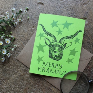 May include: A green card with a black and white illustration of a goat's head with horns and the text "Merry Krampus". The card is surrounded by five black and white stars.