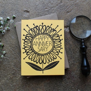 May include: A yellow card with a black and white illustration of a stylized flower. The flower has a circle in the center with the text "HAPPY ANNIVERSARY" inside.