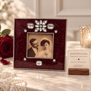 May include: A square burgundy photo frame with a textured finish, decorated with clear crystal accents and a beaded border. The frame holds a vintage-style black and white photograph of a couple. A small sign reads: Jeweled Photo Frame.
