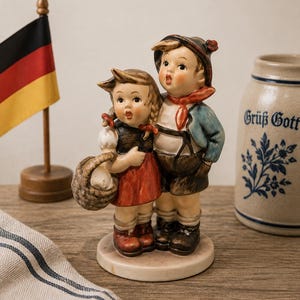 Puede incluir: Una figura de cerámica de una niña y un niño de pie juntos. La niña lleva una falda roja, un chaleco oscuro y lleva una cesta tejida. El niño lleva una chaqueta azul, pantalones cortos marrones y un pañuelo rojo al cuello.