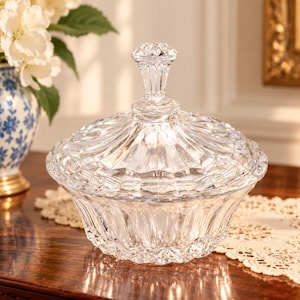 May include: A clear glass candy dish with a matching lid, featuring a faceted, diamond-cut pattern on the exterior. The bowl has a wide, rounded base with vertical fluting, and the lid is topped with a decorative, multi-faceted glass finial.