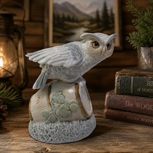 May include: A porcelain owl figurine perched on a small barrel with embossed leaf details. The owl is light blue and white with detailed feather textures and large dark eyes. The barrel is cream-colored with brown trim and sits on a textured base.