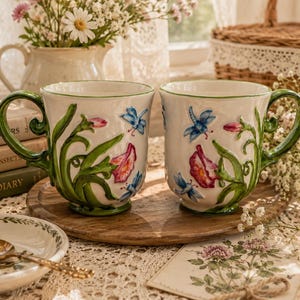 May include: Two white ceramic mugs featuring raised, hand-painted designs of green vines, pink flowers, and blue dragonflies. Each mug has a curved green handle and a flared rim, showcasing a detailed, three-dimensional botanical motif.
