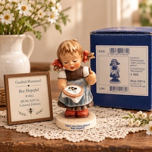 Puede incluir: Una figura Goebel Hummel titulada Bee Hopeful #1662, que muestra a una niña con coletas sosteniendo un bastidor de bordado con un diseño de abeja. La base tiene la inscripción Hab's von Oma gelernt. Incluye un letrero y una caja azul.