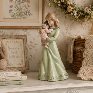 May include: A ceramic figurine of a woman in a long green dress holding a baby. The figurine is on a white surface with a framed print, a stuffed bunny, and books. The text on the frame reads "You are so loved little one."