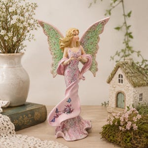 May include: A standing fairy figurine with blonde hair, wearing a long pink dress adorned with floral patterns and small blue butterflies. The figure features large, light green wings decorated with pink accents and embedded sparkling rhinestones.