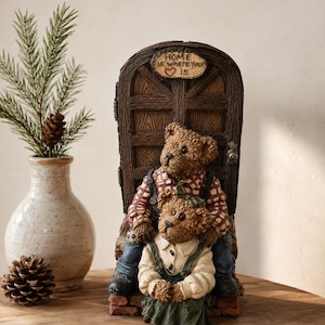 May include: A resin figurine featuring two brown teddy bears sitting in front of a dark brown arched door. The top of the door has a plaque that reads "HOME is where your heart is" with a red heart. The bears wear plaid and solid clothing.