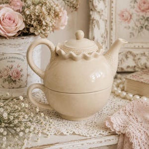 Può includere: Un set da tè in ceramica color crema composto da una teiera impilata sopra una tazza abbinata. La teiera presenta un bordo arricciato attorno al coperchio, un manico testurizzato e un beccuccio testurizzato. La tazza ha un corpo arrotondato e un manico.