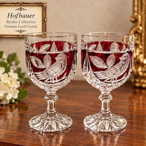 May include: Two Hofbauer Byrdes Collection German lead crystal wine glasses featuring a deep red color with clear cut glass bird and leaf motifs. The stems are faceted and the bases have a starburst pattern. A sign reads Hofbauer Byrdes Collection.