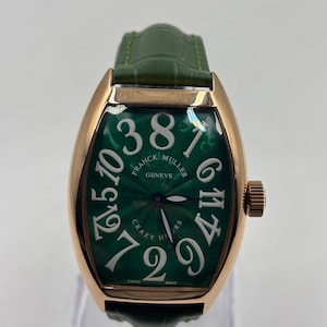 Könnte beinhalten: Eine Luxus-Armbanduhr mit grünem Zifferblatt und goldfarbenem Gehäuse. Die Uhr hat große weiße Ziffern und die Aufschrift "FRANCK MULLER GENEVE" und "CRAZY HOURS". Das Armband ist aus grünem Leder.