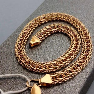 May include: A gold-toned chain necklace featuring a complex, woven, circular link pattern. The chain is constructed from interconnected metal loops that create a textured, mesh-like appearance. It includes two polished, triangular-shaped end caps.