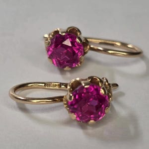 May include: A pair of gold earrings featuring vibrant pink gemstones. The gemstones are round and set in a decorative, scalloped setting. The earrings have a simple hook closure.