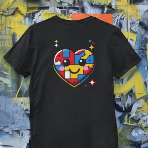 May include: Black t-shirt featuring a colorful heart-shaped graphic. The heart design is composed of puzzle-like pieces in red, yellow, blue, and pink, with a smiling face. The shirt is hanging on a metal hanger.