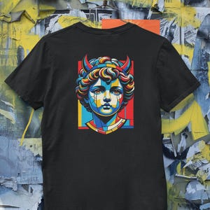 Puede incluir: Camiseta negra con una colorida ilustración de una figura similar a una estatua con cuernos. La obra de arte utiliza un estilo geométrico con azules, rojos, amarillos y naranjas vibrantes. El diseño está centrado en la camiseta.