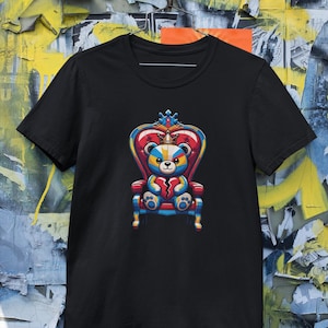Puede incluir: Camiseta negra con un gráfico colorido de un oso de peluche con corona sentado en un trono. El oso tiene un diseño de corazón roto. El fondo es una colorida pared de graffiti.