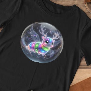 Puede incluir: Camiseta negra con una criatura caprichosa y colorida con cuernos y alas dentro de una burbuja. También se muestran pantalones cortos verde oliva con un estampado de cocodrilo, sandalias marrones y un cojín marrón con forma de perro.