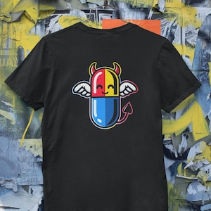 May include: Black t-shirt featuring a colorful cartoon pill design. The pill has red, yellow, and blue sections, with wings, horns, and a tail. The design is a playful take on a devilish character.